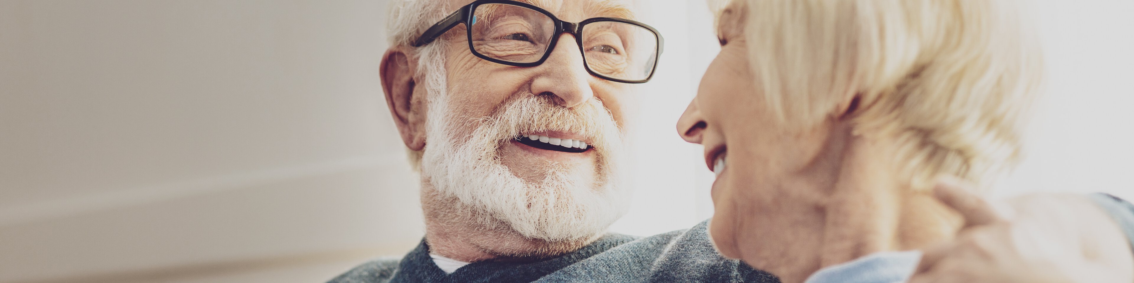 Ein Senior mit Brille sitzt neben einer Seniorin, umarmt sie und beide lächeln sich an. | © Fotolia 210582403 - Adobe Stock - Zinkevych